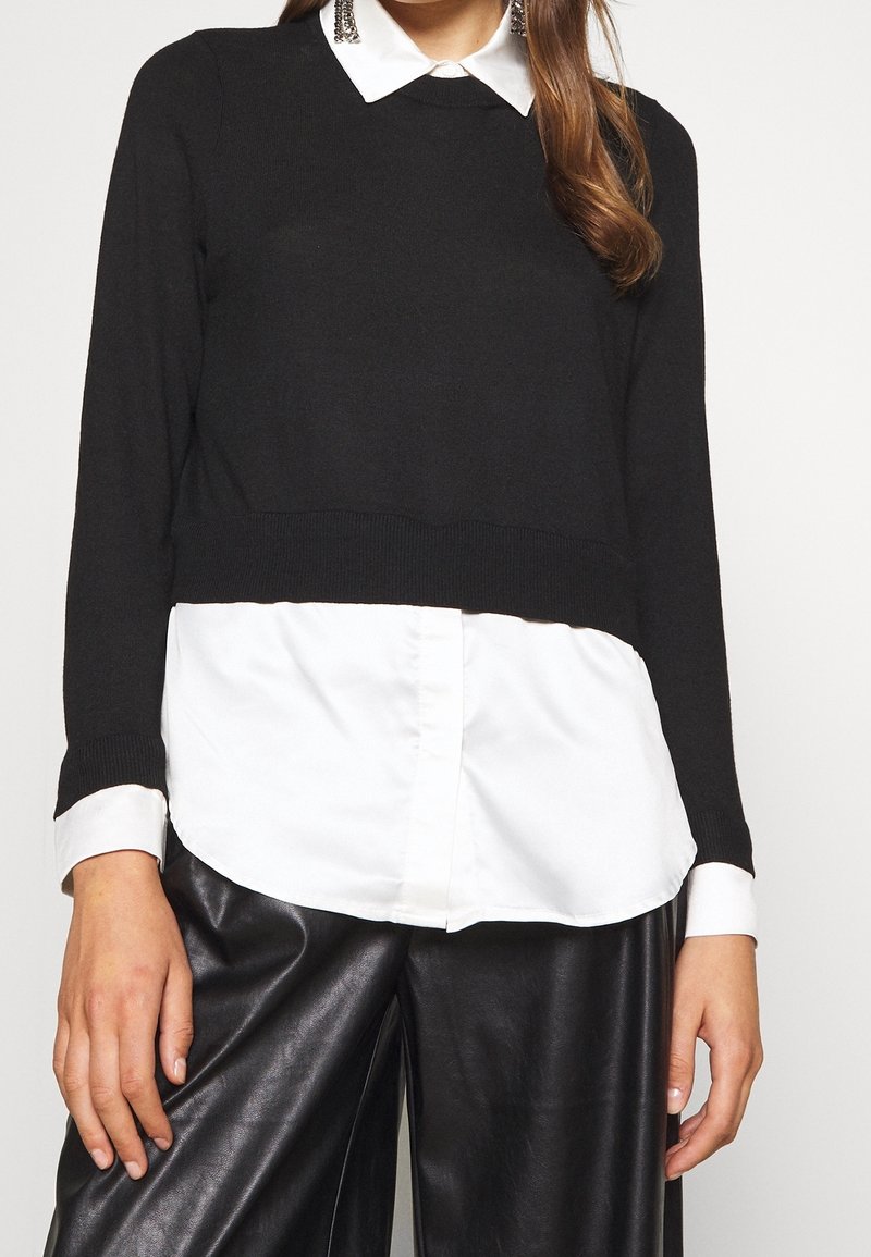 Donna che indossa un maglione corto nero sopra una camicia bianca, con pantaloni in pelle nera. Sono visibili anche orecchini pendenti e capelli sciolti.