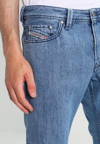 Light blue denim jeans with a straight cut, featuring a front pocket with a logo tag and metal button details. Textured fabric with slight fading.