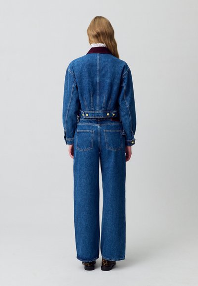 Tuta in denim con corpino aderente, gambe larghe e vita con cintura. Presenta un colletto scuro e tasche laterali con accessori gialli a contrasto.