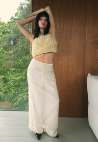 Top corto arricciato di crema abbinato a pantaloni larghi di colore chiaro. Il modello è in piedi con le braccia sollevate, mettendo in mostra un design elegante e tacchi a spillo neri.
