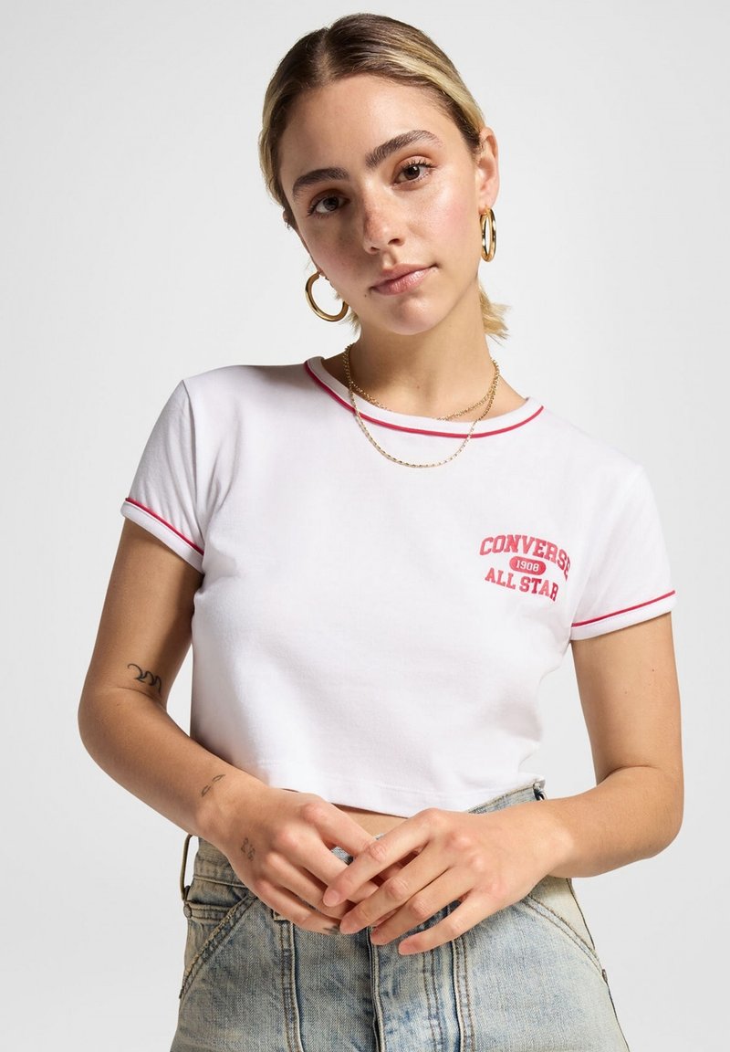 Joven mujer con pelo rubio que lleva una camiseta blanca corta con ribete rojo y el logo "Converse All Star", aretes de aro dorados y pantalones de mezclilla.