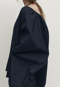 Marinblå topp med breda, flödande ärmar och öppen halsringning. Tillverkad av ett mjukt tyg med löst passform, med subtila rynkor vid axlarna.