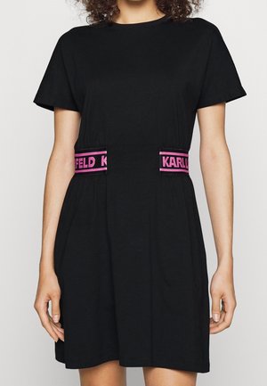 Frau trägt ein schwarzes kurzärmeliges Kleid mit pinken und schwarzen Taillenbändern, auf denen der Text "KARL LAGERFELD" in Blockbuchstaben steht.