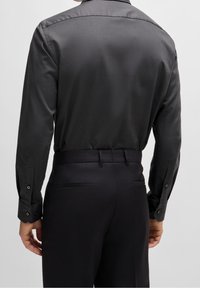 Dunkelgraues Langarmhemd mit glatter Textur, mit Knopfmanschetten und Cutaway-Kragen; kombiniert mit maßgeschneiderten schwarzen Hosen mit Gesäßtaschen.