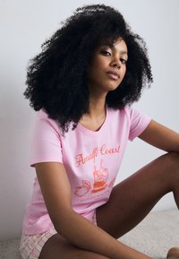T-shirt de algodão rosa com um design gráfico laranja de uma bebida com acentos cítricos, complementado por mangas curtas e um corte descontraído.