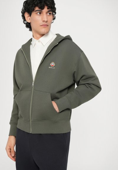 Joven con una sudadera con cremallera en color verde oliva con el logo de Bally, camisa blanca debajo y pantalones negros, mirando hacia un lado.
