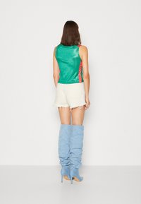 Top verde sem mangas com detalhes laterais vermelhos, combinado com calções brancos desfiados e botas azuis até ao joelho. Textura suave, design ajustado.