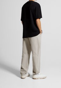 Musta puuvillapaita lyhyillä hihoilla, yhdistettynä vaaleanbeigeihin, löysiin housuihin. Mallilla on jalassa valkoiset lenkkarit.