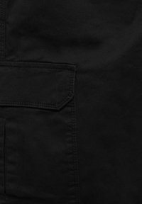 Schwarzer Stoff mit matter Oberfläche, ausgestattet mit einer aufgesetzten Tasche mit scharfen Winkeln und sichtbaren Nähten. Glatte Textur, keine Muster oder Beschläge.