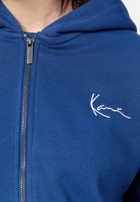 Felpa blu con zip realizzata in tessuto morbido. Presenta una zip frontale e un logo bianco ricamato sul lato sinistro del petto.