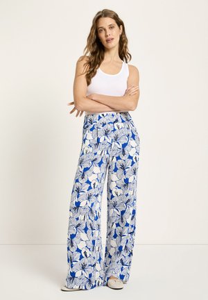 Frau steht mit verschränkten Armen, trägt ein weißes Tanktop, weite Hosen mit blau-weißem Blumenmuster und beige Ballerinas vor einfachem Hintergrund.