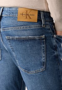 Plave traperice imaju teksturiranu obradu, lagano blijeđenje i ukrasne šavove. Na leđima se vidi smeđa kožna oznaka s natpisom "Calvin Klein Jeans".
