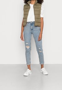 Person wearing olive green puffer vest over white eyelet short-sleeve top, ripped light blue high-rise jeans, and white sneakers, standing.