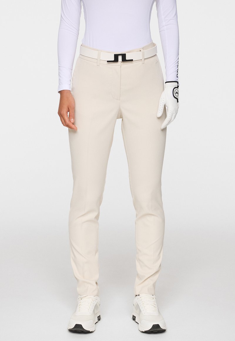 Beige golfhousut, jotka on valmistettu joustavasta kankaasta, joissa on slim fit -malli, etutaskut, musta vyö ja valkoiset urheilukengät. Silkkinen pinta.