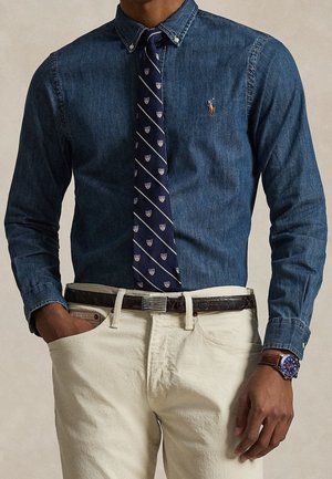 Homme portant une chemise en jean bleu boutonnée, une cravate bleu marine à motifs, un pantalon beige, une ceinture foncée et une montre bleue avec un bracelet en cuir marron.