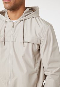 Ljusbeige huvjacka gjord av ett glatt, vattenavvisande material. Har en framzipper, dragsko på huvan och subtil logotypdetalj.