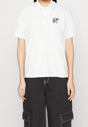 Personne portant un polo blanc avec un crocodile coloré sur un logo de vélo, associé à un pantalon cargo noir avec des coutures blanches.