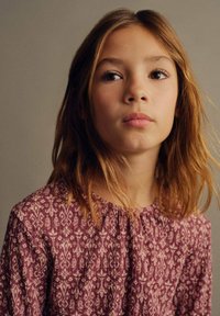 Una giovane con capelli castano medio che indossa un vestito rosa a fantasia con scollatura arricciata e motivi floreali intricati.