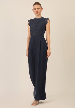 Jumpsuit marine avec un haut en dentelle doté de manches courtes, d'un col haut et de pantalons larges et fluides avec des plis. Sandales argentées simples.