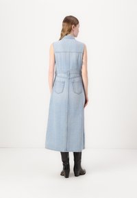 Ermeløs denimkjole i lyseblå, med belte i midjen, baklommer og en lang, flytende silhuett, kombinert med blanke svarte støvler.