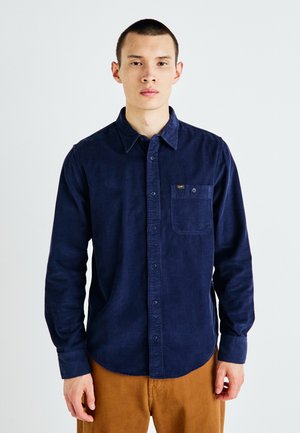 Marineblauwe ribfluwelen overhemd met lange mouwen, borstzak en gouden logo-accent; gestyled met kaki broeken.
