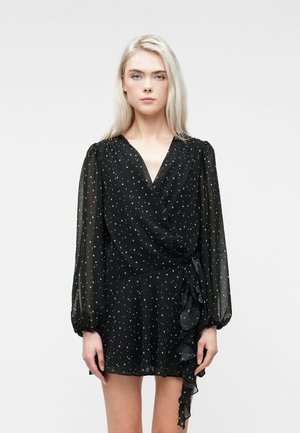 Młoda kobieta z długimi blond włosami ubrana w czarną sukienkę w stylu wrap z przezroczystymi długimi rękawami i małym białym wzorem w gwiazdki.