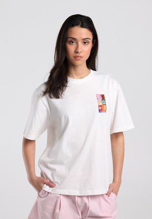 Young woman with dark hair wears white oversized T-shirt with small colorful abstract design on chest and light pink pants, hands in pockets.
