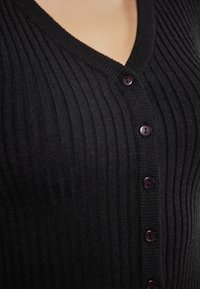 Černý žebrovaný cardigan s výstřihem do V, zdobený tmavými knoflíky na přední straně. Měkký materiál s viditelnou texturou a jemnými švy.