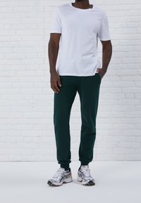 Man i vit t-shirt, mörkgröna joggingbyxor och vita löparskor står mot en vit tegelvägg.