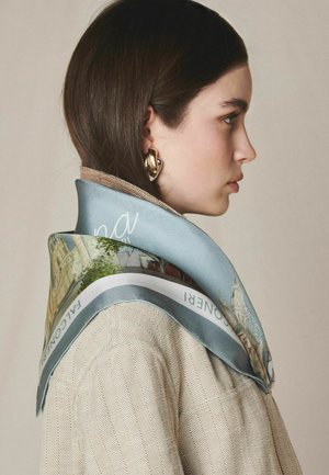 Kobieta na profil ma na sobie beżowy płaszcz w jodełkę, jasnoniebieski szalik z motywem miejskim oraz duże złote kolczyki koła na gładkim tle.