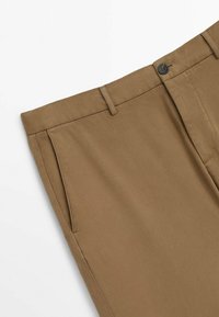 Zbliżenie na beżowe spodnie, pokazujące szlufki na pasek, boczną kieszeń oraz zapięcie na guzik w talii.