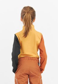 Väriesteröity takki, jossa on keltaisia, oransseja ja mustia paneeleja; korkea kaulus; sileä pinta; yhdistetty oransseihin housuihin, joissa on joustava vyötärö.