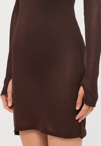 Rjava rebrasta pletena obleka z dolgimi rokavi in odprtinami za prste, prilegajoča se oblika in raztrgani robovi.