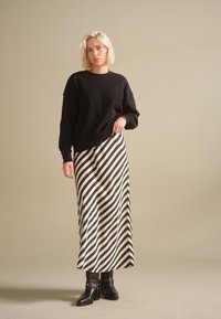 Kobieta o blond włosach ubrana w czarną bluzę, czarno-białą spódnicę w ukośne paski oraz czarne, ćwiekowane botki, stojąca na jednolitym tle.