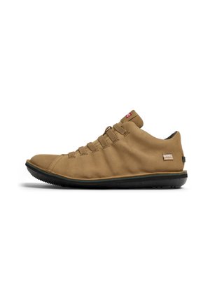 Beige Outdoor-Schuhe mit einem weichen Wildleder-Obermaterial, elastischen Schnürsenkeln, flachem Profil und einer schwarzen Gummisohle. Auf der Seite befindet sich ein kleines Logo-Tag.