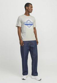 Hellgrau kurzärmeliges T-Shirt mit "JACK&JONES" Grafik in Blau und Weiß, kombiniert mit dunklen Jeans und weißen Sneakern.