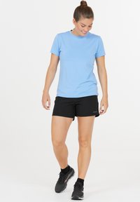 Ljusblå kortärmad träningsskjorta, kombinerad med svarta shorts. Modell som bär svarta träningsskor, stående mot en helt vit bakgrund.