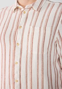 Camicia abbottonata realizzata in tessuto testurizzato con strisce verticali bianche e rosse. Presenta una tasca sul petto e bottoni beige.