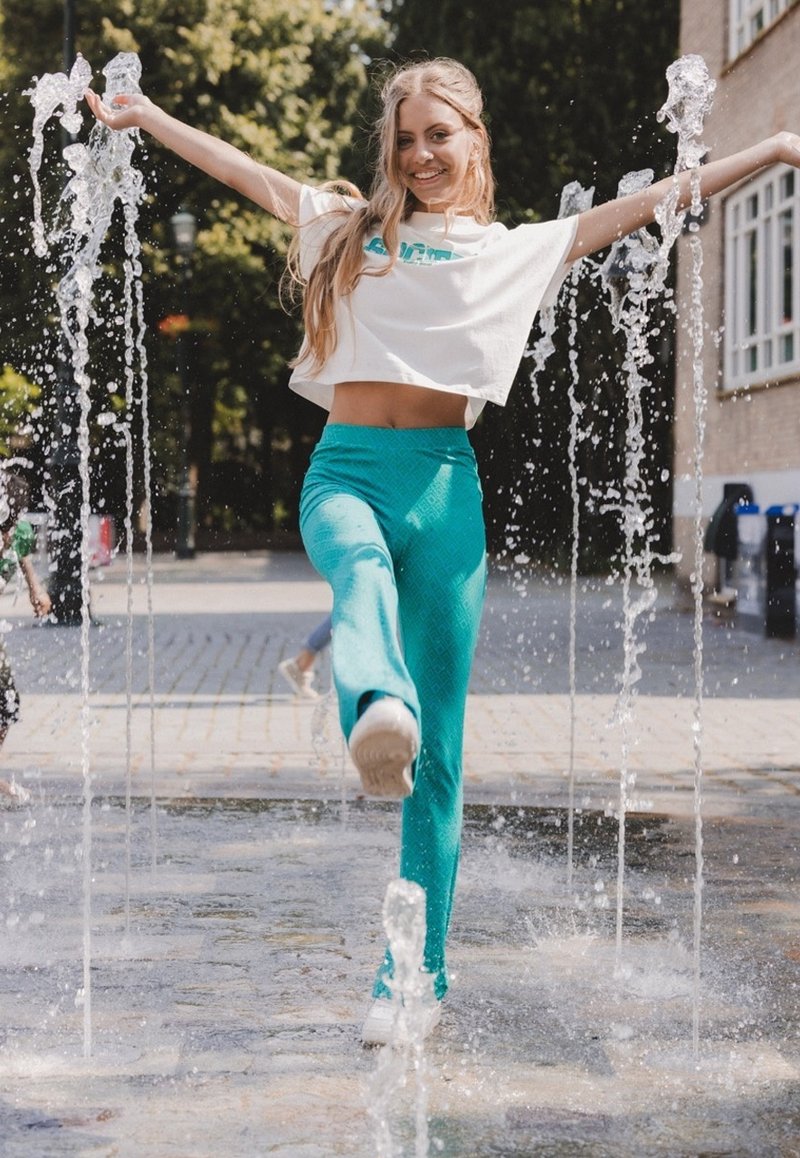 Een persoon in een kort wit T-shirt en teal-gepatenteerde broek poseert speels in een fontein, terwijl het water om hen heen spettert.