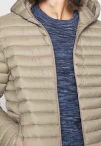 Beige gewatteerde jas met horizontale doorgestikte textuur en capuchon, gedragen over een blauwe gebreide trui met structuur. Voorzien van een ritssluiting aan de voorkant.