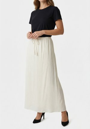 Mujer con top negro de manga corta, falda larga blanca texturizada con cintura ajustable y zapatos de tacón negro, de pie frente a un fondo blanco.