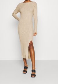 Robe moulante en maille côtelée beige clair avec manches longues et fente haute sur la cuisse, associée à des sandales plates noires.