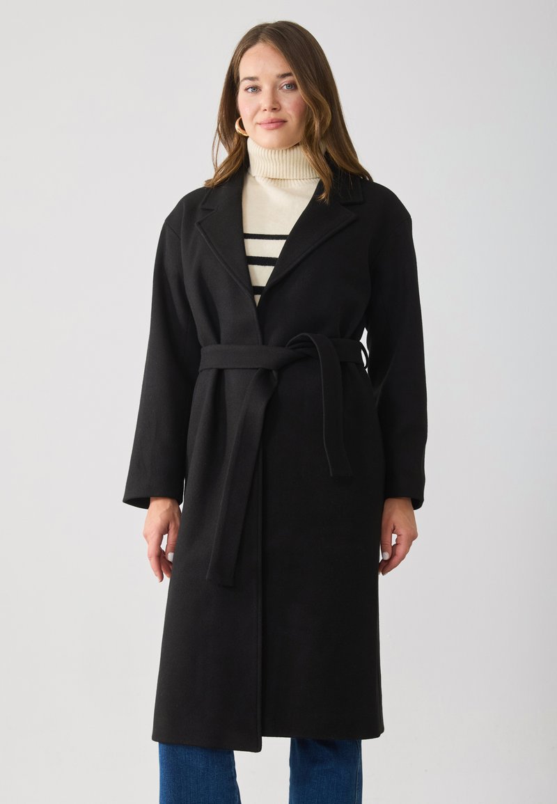 Cappotto nero con cintura in vita, colletto a pistagna, maniche lunghe e texture morbida, indossato su un dolcevita crema con righe nere. Silhouette aderente.