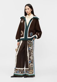 Mladá žena v tmavohnedých vzorovaných širokých nohaviciach a nadrozmernom hnedom svetri s dekoratívnymi vzormi, stojaca s rukou v vrecku.