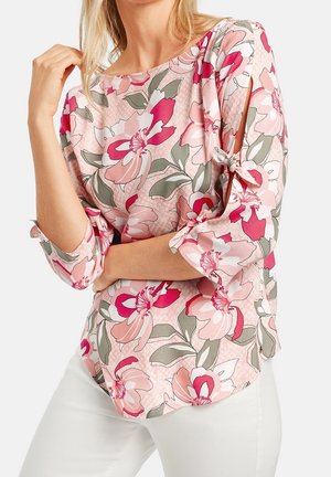 Femme portant un chemisier rose à fleurs avec des manches trois-quarts attachées aux poignets et un pantalon blanc sur un fond uni.