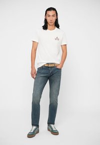 Bílý bavlněný tričko s logem hory, v kombinaci s modrými džínami. Uživatel má na sobě béžový pásek a zelené tenisky s bílými detaily.