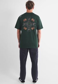 T-shirt en coton vert foncé avec un motif graphique d'insectes et de fleurs dans le dos. Coupe décontractée, manches courtes, col rond.