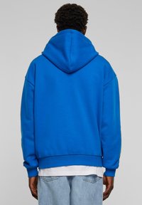 Sininen huppari, joka on valmistettu pehmeästä kankaasta. Ominaisuuksina rento leikkaus ja ribbaatut hihansuut. Päällä valkoinen paita ja näkyvät vaaleansiniset farkut.