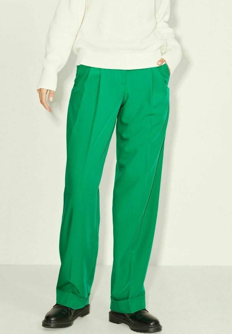 Personne portant un pantalon large vert vif, un pull en maille blanc et des chaussures en cuir noires, debout avec une main dans la poche.