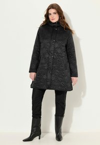 Svart quiltad kappa med hög krage, tryckknappar, sidofickor och ett texturerat vågmönster. Matchad med svarta snäva jeans och höga stövlar.